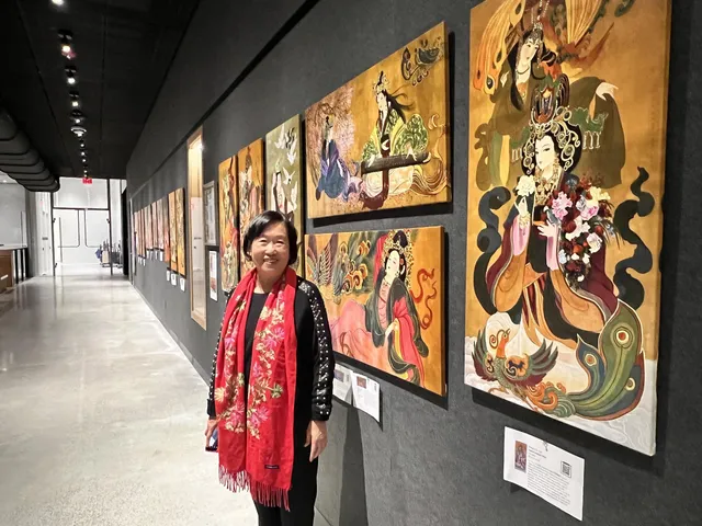 Xiang Li in a red scarf beside a row of large Chinese empress canvases along a corridor of the Prior Performing Arts Center at Holy Cross