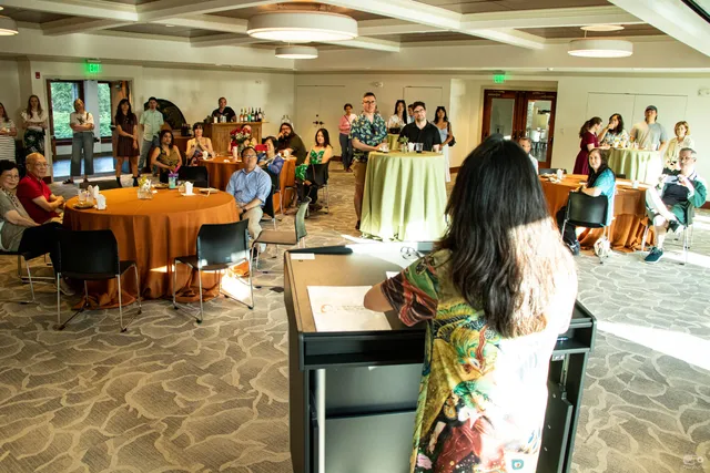 Fei Wu at a lectern speaking to guests seated at round tables in a reception room