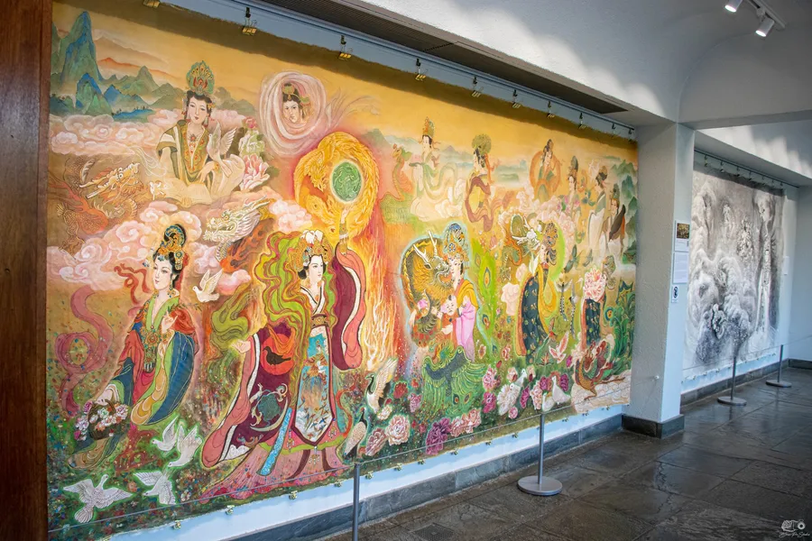 A wall-length silk painting of Chinese empresses in jewel colors hanging in a gallery at the New England Botanic Garden, an ink mural beyond it