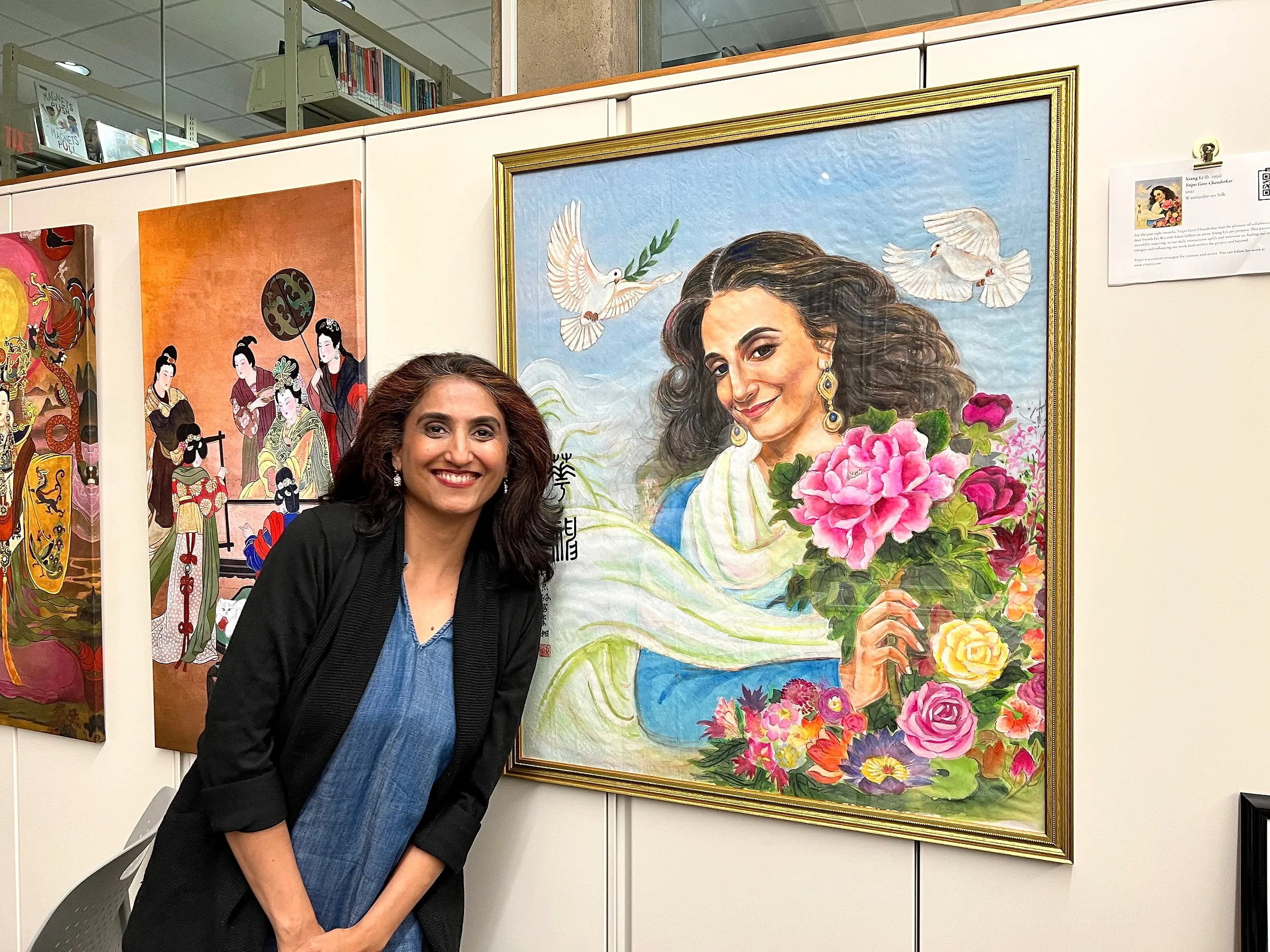 Tripti Gore Chandorkar smiling beside her framed portrait on a library wall, two of Xiang Li’s canvases hanging to the left