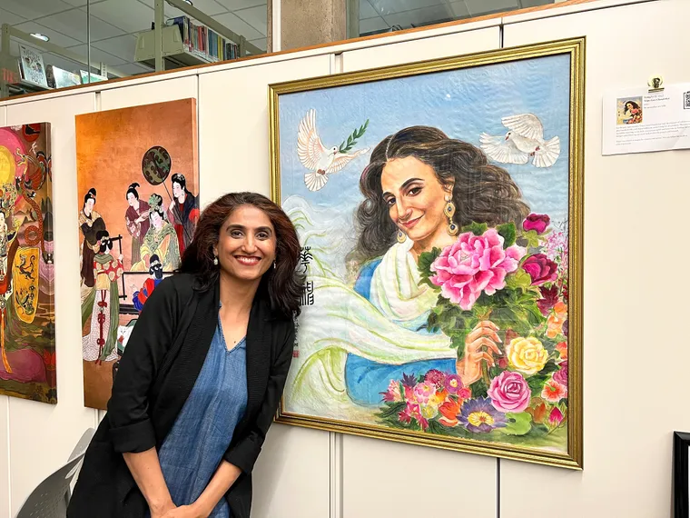 Tripti Gore Chandorkar smiling beside her framed portrait on a library wall, two of Xiang Li’s canvases hanging to the left