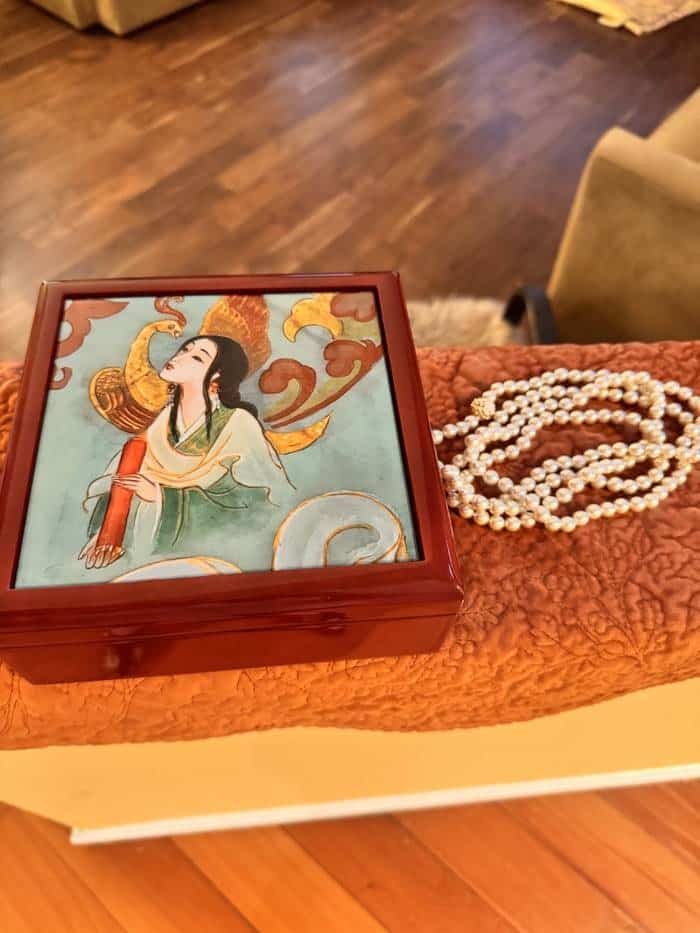 The closed jewelry box on a coral quilted bench beside a long strand of pearls.