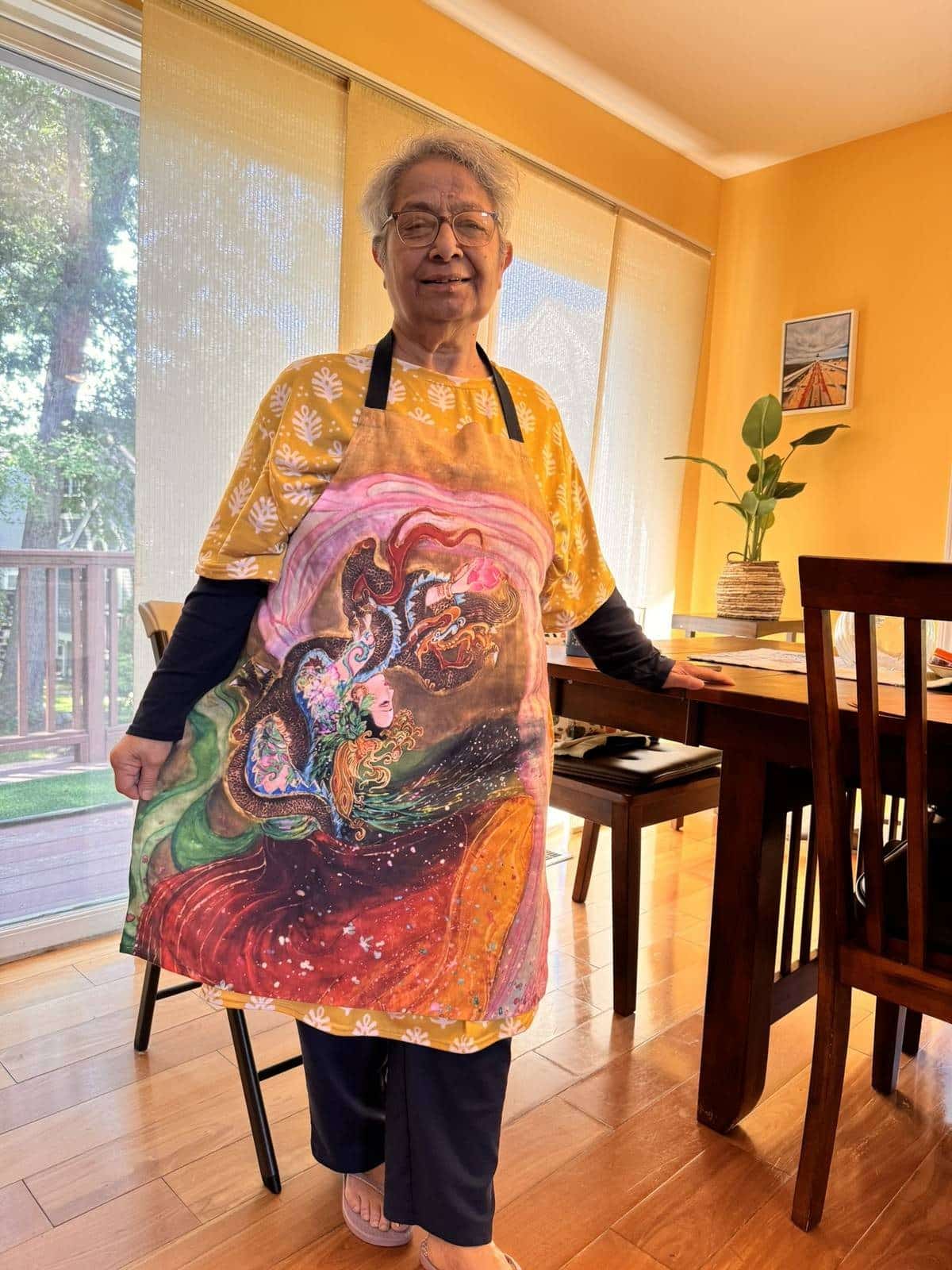 A woman standing in a sunlit dining room wearing the apron printed with the dancing Empress Li Lingrong painting.