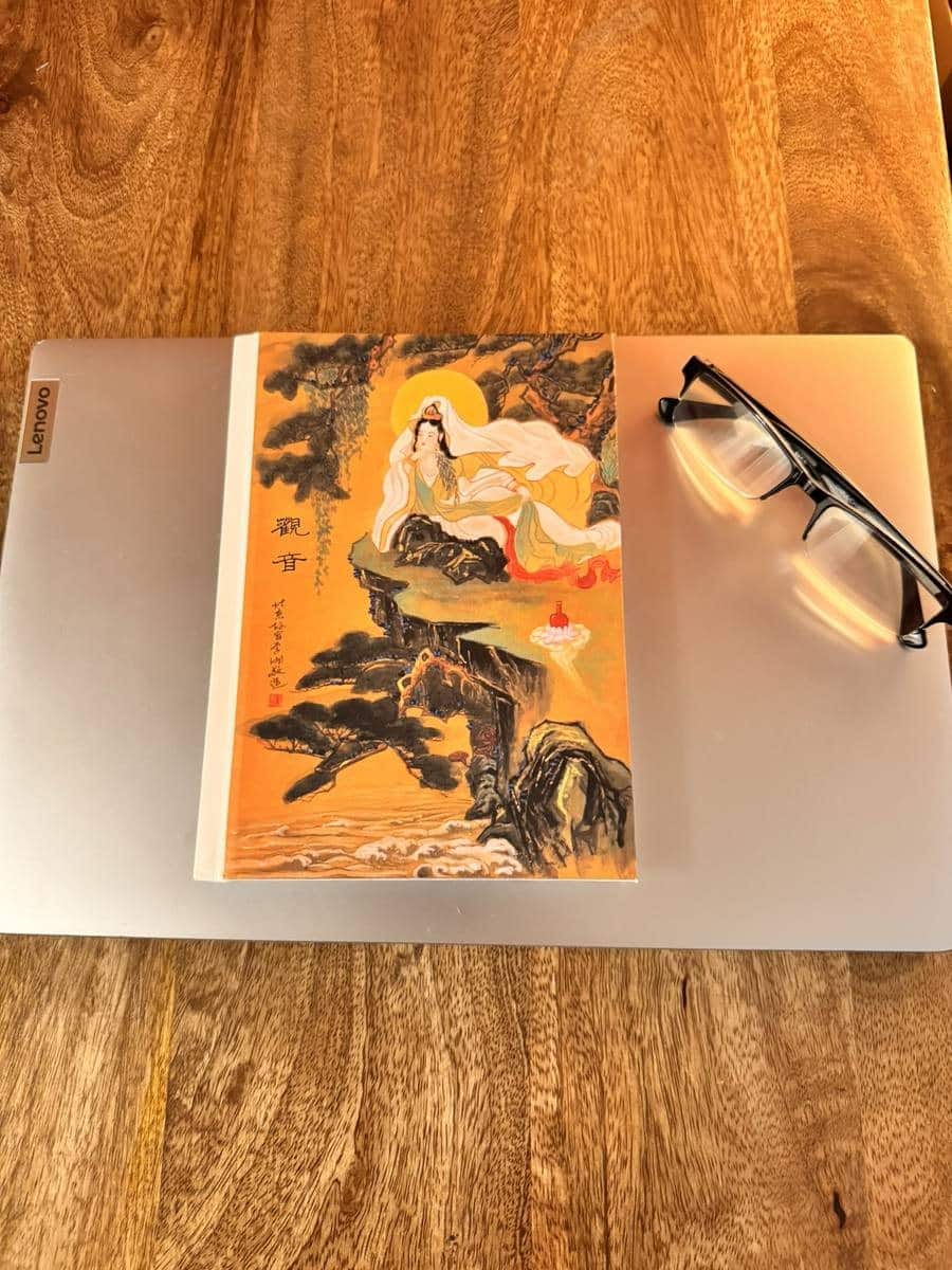The Guanyin journal lying closed on a laptop beside a pair of reading glasses on a wooden table.