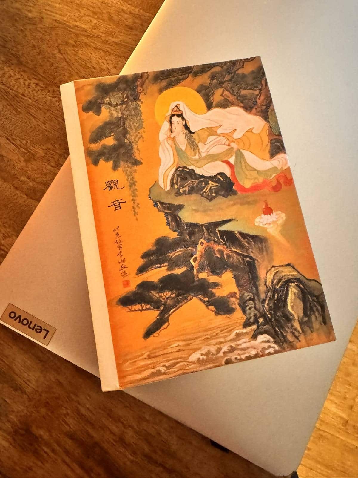 Hardbound journal with a painting of Guanyin on the cover, resting at an angle on a closed silver laptop.